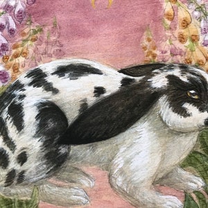 May include: A watercolor painting of a black and white spotted rabbit with long ears, positioned in a garden setting with tall purple and yellow foxglove flowers and a small gold crescent moon centered in the dusty pink sky above the animal.