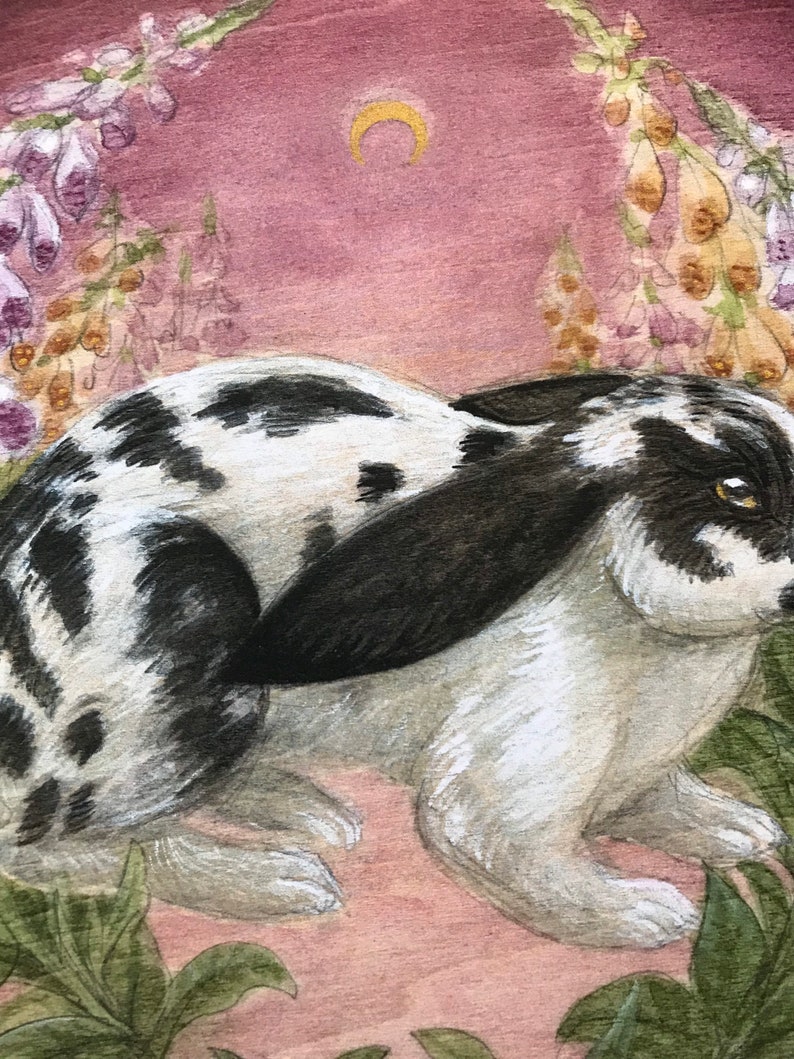 May include: A watercolor painting of a black and white spotted rabbit with long ears, positioned in a garden setting with tall purple and yellow foxglove flowers and a small gold crescent moon centered in the dusty pink sky above the animal.