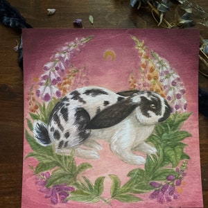 May include: A square art print on wood featuring a white rabbit with black spots and long ears. The rabbit is surrounded by green leaves and tall purple and white foxglove flowers against a deep pink background with a small yellow crescent moon.