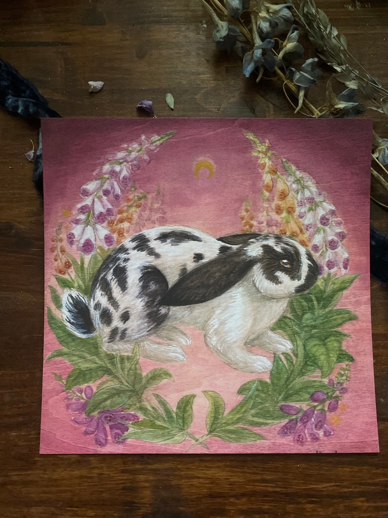 May include: A square art print on wood featuring a white rabbit with black spots and long ears. The rabbit is surrounded by green leaves and tall purple and white foxglove flowers against a deep pink background with a small yellow crescent moon.