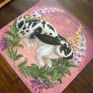 May include: A square art print featuring a black and white spotted rabbit centered within a circular wreath of green leaves and purple foxglove flowers against a pink background with a small yellow crescent moon in the upper right corner.