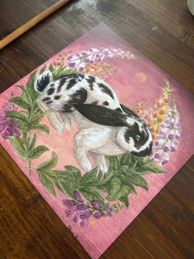 May include: A square art print featuring a black and white spotted rabbit centered within a circular wreath of green leaves and purple foxglove flowers against a pink background with a small yellow crescent moon in the upper right corner.
