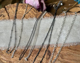 May include: An assortment of silver and black necklaces, presented on a wooden surface. The necklaces display different chain designs and small pendant features, reflecting a minimalist jewellery style.