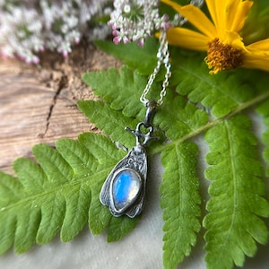 May include: A silver pendant necklace featuring a moth design with a pear-shaped, iridescent moonstone. The pendant hangs from a delicate chain and rests on a green fern leaf, with a yellow flower and white flowers in the background.