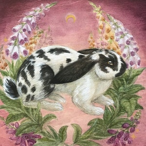 May include: A painting of a black and white spotted rabbit centered among purple and yellow foxglove flowers. A small gold crescent moon is positioned above the rabbit against a dark pink background. The artist signature is in the bottom right.