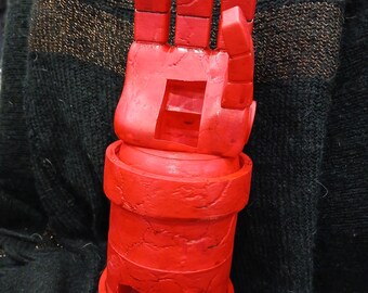 May include: A bright red, articulated hand prop, likely for cosplay or display. The hand is made of a solid material, with visible joints and a cylindrical base. The fingers are straight and square, and the overall design is stylized.