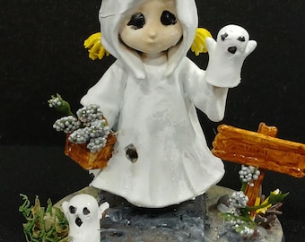 May include: A handcrafted Halloween figurine of a ghost child in a white hooded robe, holding a small ghost and a basket of flowers. The figurine is on a stone base with a wooden sign.