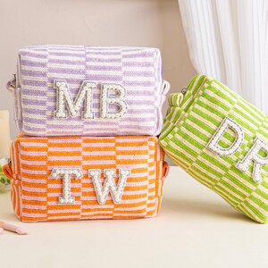 Könnte beinhalten: Drei personalisierte Kosmetiktaschen in einem Stapel. Die obere Tasche ist lila und weiß kariert, mit den Initialen "MB". Die mittlere Tasche ist orange und weiß kariert, mit den Initialen "TW". Die untere Tasche ist grün und weiß gestreift, mit den Initialen "DR".