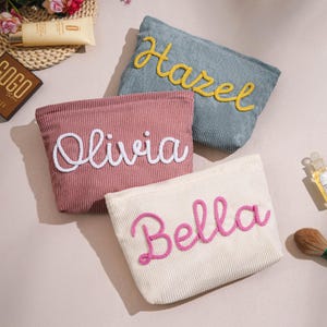 Op de afbeelding: Vier corduroy make-up tasjes in roze, crème en blauwgroen. Elke tas heeft een naam geborduurd in een contrasterende kleur: "Hazel" in geel, "Olivia" in wit en "Bella" in roze. De tasjes zijn rechthoekig met een opening aan de bovenkant.