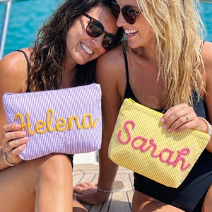 May include: Two women holding personalized seersucker pouches. One pouch is lavender with "Helena" embroidered in yellow, the other is yellow with "Sarah" in pink. Both pouches have a zipper closure and are on a boat.