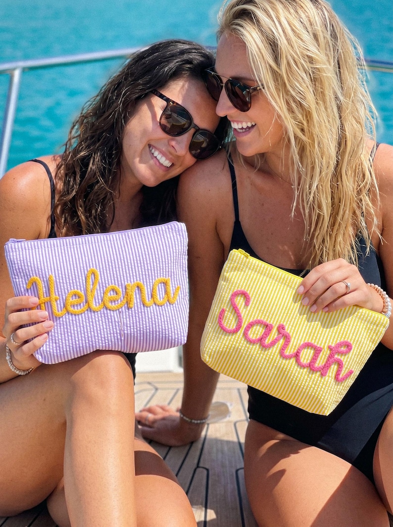 May include: Two women holding personalized seersucker pouches. One pouch is lavender with "Helena" embroidered in yellow, the other is yellow with "Sarah" in pink. Both pouches have a zipper closure and are on a boat.