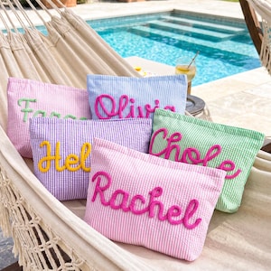 Puede incluir: Bolsas de cosméticos de seersucker en colores pastel, como rosa, morado, azul y verde. Cada bolsa tiene un nombre bordado en un color contrastante. Las bolsas están dispuestas en una hamaca cerca de una piscina.