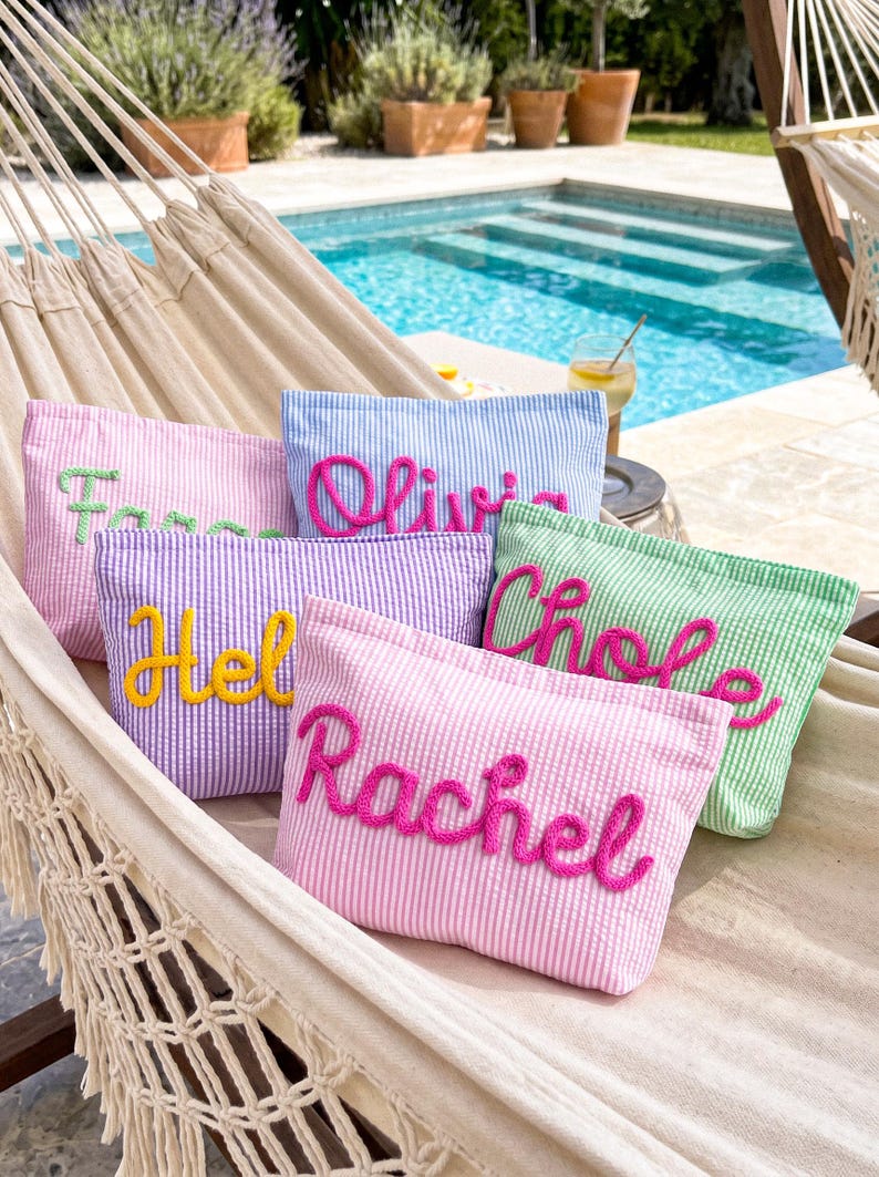 May include: A collection of seersucker cosmetic bags in various pastel colors, including pink, purple, blue, and green. Each bag features a name embroidered in a contrasting color. The bags are arranged on a hammock near a pool.