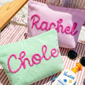 May include: Two seersucker pouches, one pink and one green, each with a name embroidered in bright pink. The pink pouch says "Rachel", and the green pouch says "Chloe". Sunglasses and sunscreen are also visible.