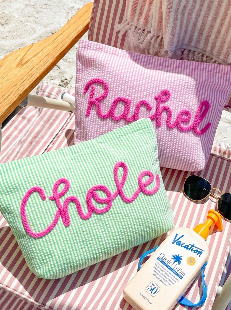 May include: Two seersucker pouches, one pink and one green, each with a name embroidered in bright pink. The pink pouch says "Rachel", and the green pouch says "Chloe". Sunglasses and sunscreen are also visible.