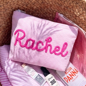 May include: Pink and white striped cosmetic bag with the name "Rachel" embroidered in bright pink thread. The bag is inside a woven basket, with beauty products and a water bottle visible.