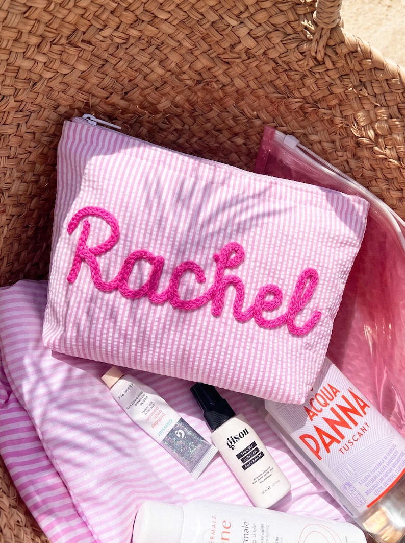 May include: Pink and white striped cosmetic bag with the name "Rachel" embroidered in bright pink thread. The bag is inside a woven basket, with beauty products and a water bottle visible.