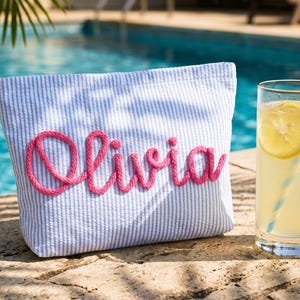 May include: A blue and white striped pouch with the name "Olivia" embroidered in pink. A glass of lemonade with a lemon slice and a striped straw sits next to the pouch, with a pool in the background.
