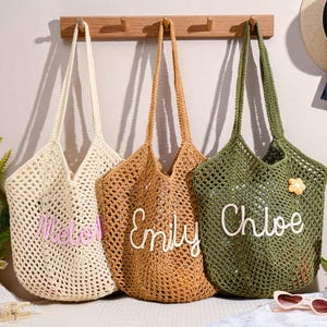 Puede incluir: Tres bolsos de mano de crochet en crema, marrón y verde oliva cuelgan de un perchero de madera. Cada bolso tiene un nombre bordado: "Madel", "Emily" y "Chloe". También se ve un sombrero de paja con una cinta negra y gafas de sol.