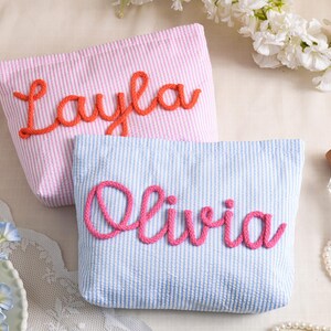 May include: Two seersucker cosmetic bags, one pink with "Layla" in orange script, the other blue with "Olivia" in pink script. Both bags have a striped pattern and are displayed with perfume and flowers.