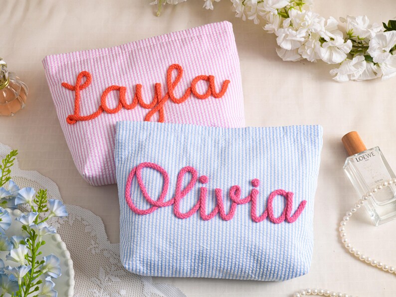 May include: Two seersucker cosmetic bags, one pink with "Layla" in orange script, the other blue with "Olivia" in pink script. Both bags have a striped pattern and are displayed with perfume and flowers.