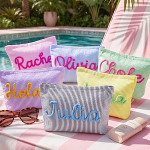May include: A collection of personalized seersucker cosmetic bags in various colors, each embroidered with a name in a contrasting color. The bags are arranged on a striped surface with sunglasses and sunscreen, suggesting a beach or poolside setting.