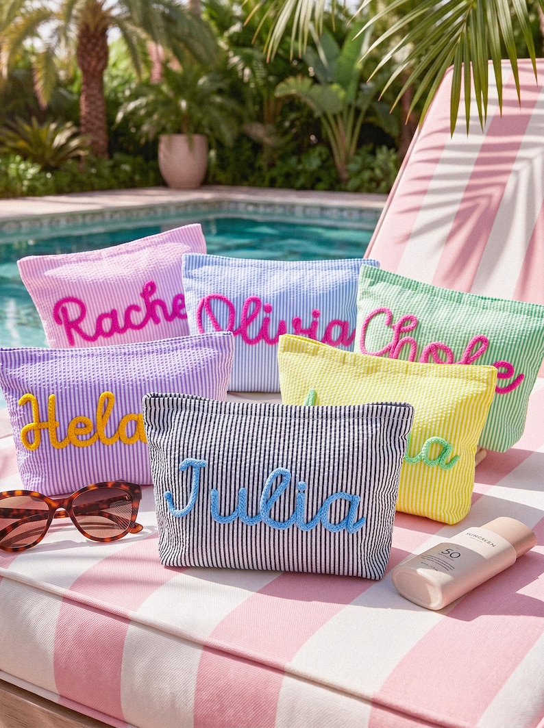 May include: A collection of personalized seersucker cosmetic bags in various colors, each embroidered with a name in a contrasting color. The bags are arranged on a striped surface with sunglasses and sunscreen, suggesting a beach or poolside setting.