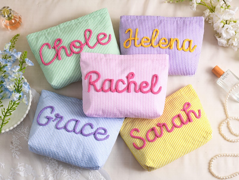 May include: Five seersucker cosmetic bags in pastel colors: green, purple, pink, blue, and yellow. Each bag has a name embroidered in a contrasting color: Chole, Helena, Rachel, Grace, and Sarah. The bags are rectangular with a zipper closure.