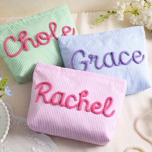 May include: Three seersucker cosmetic bags in pastel colors: green, blue, and pink. Each bag has a name embroidered in a contrasting color: "Chloe" in pink, "Grace" in purple, and "Rachel" in pink. The bags are arranged on a white surface with floral accents.