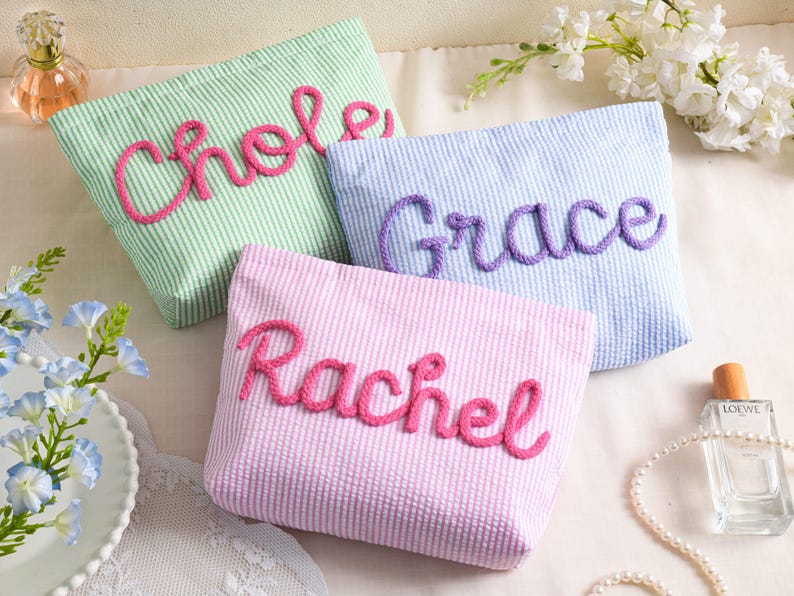 May include: Three seersucker cosmetic bags in pastel colors: green, blue, and pink. Each bag has a name embroidered in a contrasting color: "Chloe" in pink, "Grace" in purple, and "Rachel" in pink. The bags are arranged on a white surface with floral accents.