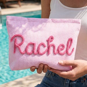 May include: Pink and white striped pouch with the name "Rachel" embroidered in pink script. The pouch is held in front of a pool, suggesting a summer or beach theme. The fabric has a textured seersucker appearance.