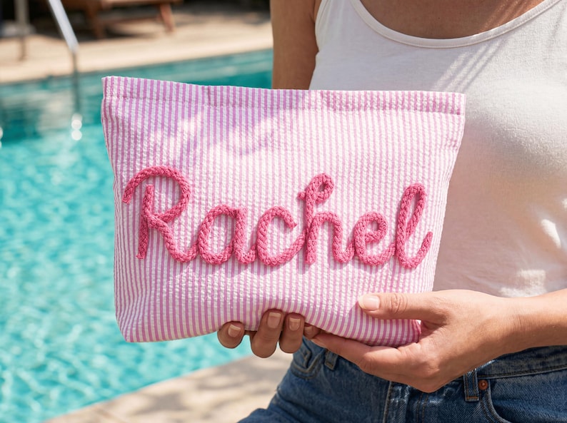 May include: Pink and white striped pouch with the name "Rachel" embroidered in pink script. The pouch is held in front of a pool, suggesting a summer or beach theme. The fabric has a textured seersucker appearance.