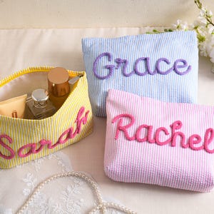 May include: Three personalized cosmetic bags in yellow, blue, and pink seersucker fabric. The bags are embroidered with the names "Sarah", "Grace", and "Rachel" in a contrasting color. The yellow bag is open, revealing makeup items.