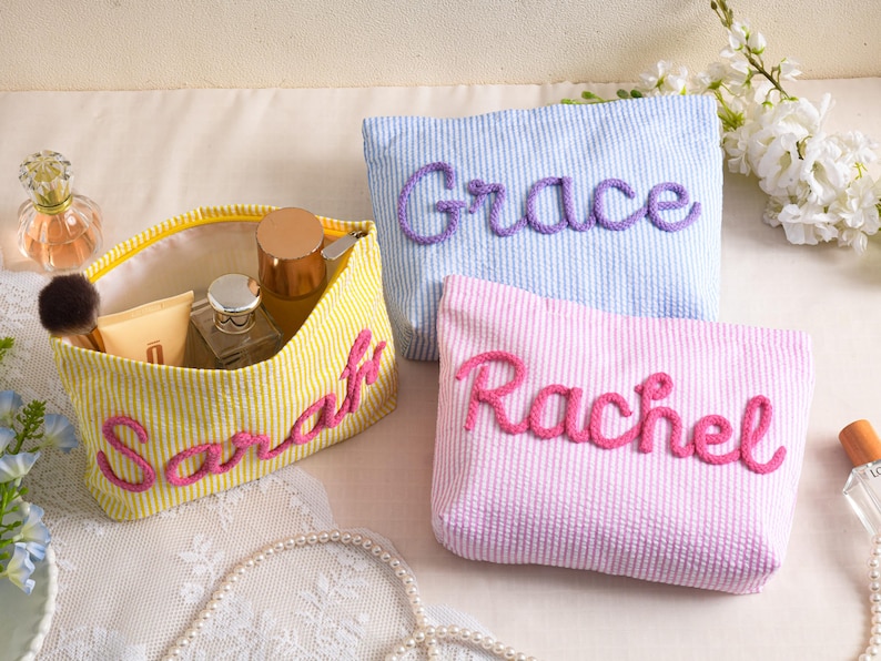 May include: Three personalized cosmetic bags in yellow, blue, and pink seersucker fabric. The bags are embroidered with the names "Sarah", "Grace", and "Rachel" in a contrasting color. The yellow bag is open, revealing makeup items.