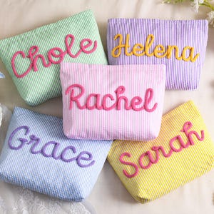 May include: Five seersucker cosmetic bags in pastel colors: green, purple, pink, blue, and yellow. Each bag has a name embroidered in a contrasting color: Chole, Helena, Rachel, Grace, and Sarah. The bags are arranged on a white surface.