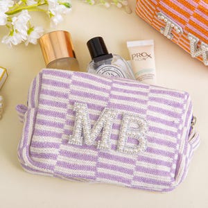 Könnte beinhalten: Eine lavendel-weiß karierte Kosmetiktasche mit den Initialen "MB" in Silber und Perlenverzierungen. Die Tasche enthält Schönheitsprodukte. Eine orange-weiß karierte Tasche mit den Initialen "LW" ist ebenfalls sichtbar.