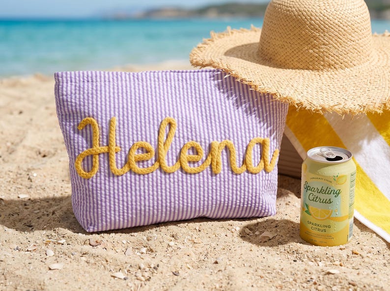 May include: A beach scene with a light purple seersucker tote bag with the name "Helena" in yellow rope lettering. A straw hat, a yellow and white striped towel, and a can of Sparkling Citrus are also visible.