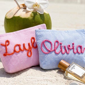 May include: Two seersucker cosmetic bags, one pink with "Layla" in orange script, the other blue with "Olivia" in pink script. A green coconut with a straw and white flower sits behind them. A bottle of perfume rests on the sand.