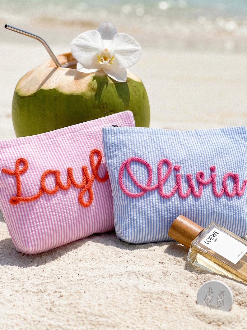 May include: Two seersucker cosmetic bags, one pink with "Layla" in orange script, the other blue with "Olivia" in pink script. A green coconut with a straw and white flower sits behind them. A bottle of perfume rests on the sand.