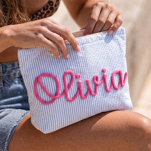May include: A blue and white striped pouch with the name "Olivia" embroidered in pink. The pouch is rectangular and has a zipper closure. The background is blurred, suggesting an outdoor setting.