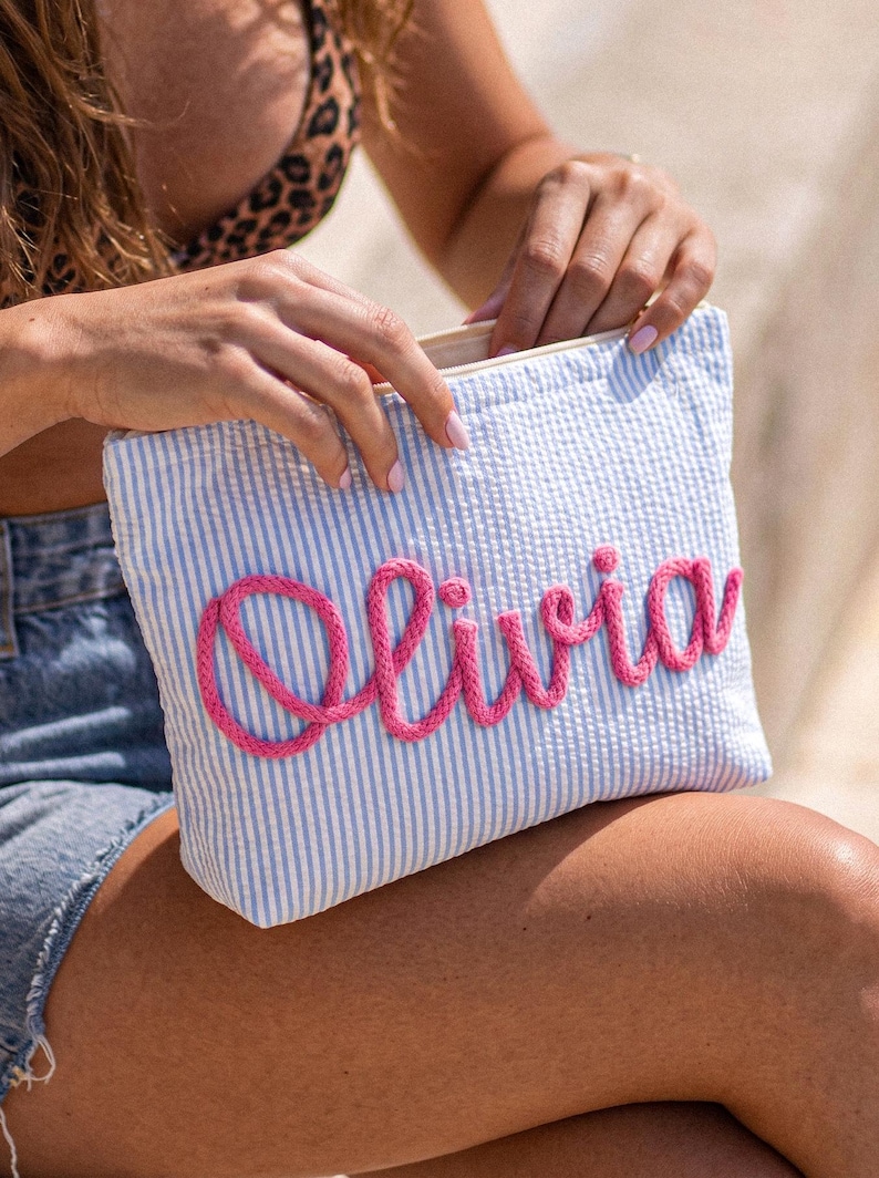 May include: A blue and white striped pouch with the name "Olivia" embroidered in pink. The pouch is rectangular and has a zipper closure. The background is blurred, suggesting an outdoor setting.
