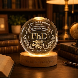 Może przedstawiać: Przezroczysta szklana kula z grawerunkiem "THE YEAR I BECAME" i "PhD", z motywem czapki absolwenta i zwoju. Kula jest podświetlana od dołu i spoczywa na drewnianej podstawie z napisem "We are so proud of you!"