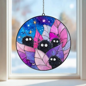 May include: Round stained glass window art featuring a whimsical design. The artwork depicts fluffy black creatures with white eyes nestled among pink and purple leaves, set against a starry blue and purple night sky. A decorative chain is attached for hanging.