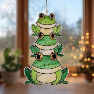 May include: A decorative hanging ornament featuring three stacked green frogs with large, gold-rimmed eyes. The frogs have white bellies and are connected by a gold string for hanging. The ornament is made of a stained glass-like material.