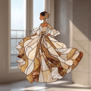 Può includere: Un ornamento in vetro colorato raffigurante una donna in un abito fluente. L'abito è un patchwork di vetro bianco, crema e marrone con accenti dorati. La donna ha i capelli scuri e indossa gioielli d'oro. L'ornamento è appeso a una catena d'oro.