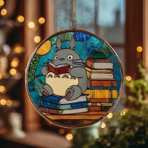 Puede incluir: Arte de vidriera con un personaje Totoro gris leyendo un libro, sentado sobre una pila de libros. La ventana redonda tiene un marco dorado y un fondo azul, verde y amarillo.