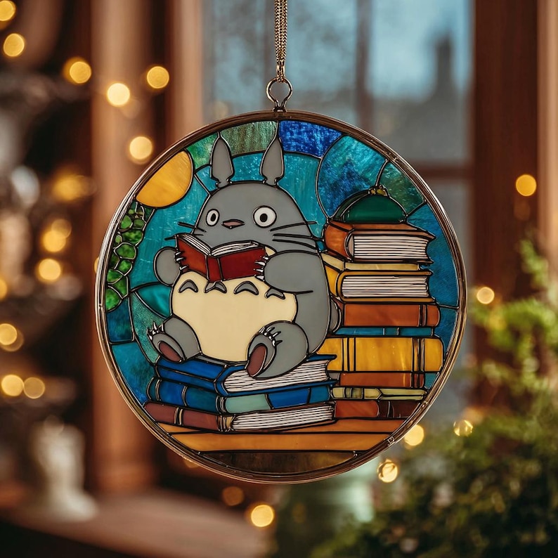 Puede incluir: Arte de vidriera con un personaje Totoro gris leyendo un libro, sentado sobre una pila de libros. La ventana redonda tiene un marco dorado y un fondo azul, verde y amarillo.
