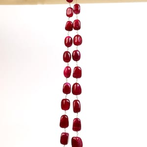 May include: A long necklace with large, polished, rectangular red gemstones strung together with silver-coloured links. The necklace hangs against a white background, with a cream-coloured fabric at the top.