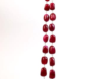 Puede incluir: Un collar largo con grandes piedras preciosas rojas rectangulares pulidas, ensartadas con eslabones plateados. El collar cuelga sobre un fondo blanco, con una tela color crema en la parte superior.