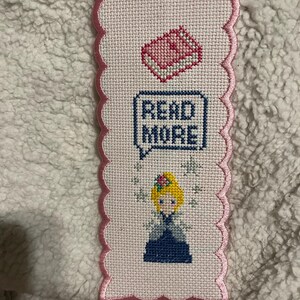 May include: Cross-stitched bookmark with a pink scalloped border. The bookmark features a pink book, the text "READ MORE" in a speech bubble, and a cartoon illustration of a blonde character. The background is white.